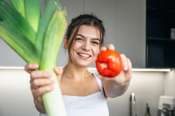 Joyeux jeune femme dans la cuisine tenant un poireau et une tomate dans ses mains, le concept de manger et de cuisiner sainement. - Photo, image
