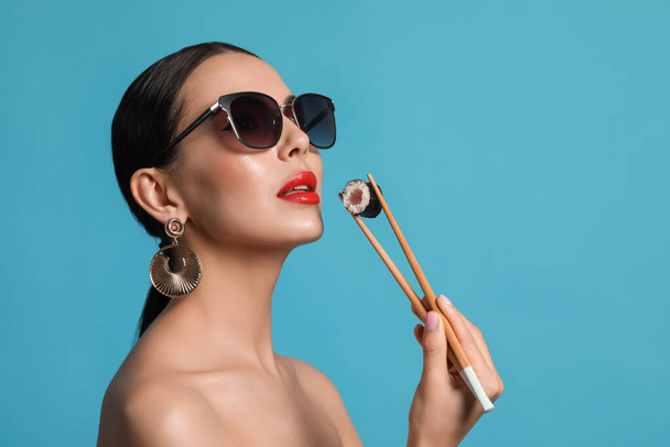 Attractive woman in fashionable sunglasses holding chopsticks with sushi against light blue background - Photo, Image