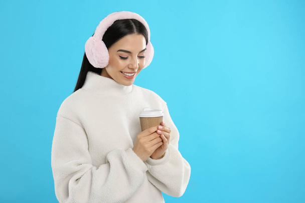 Beautiful young woman in earmuffs with cup of drink on light blue background. Space for text - Foto, imagen