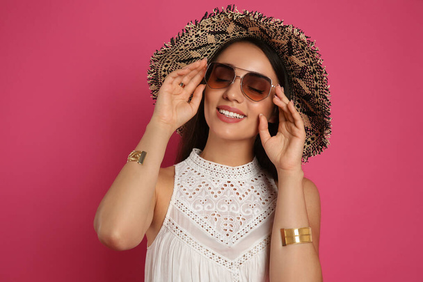 Beautiful young woman with straw hat and sunglasses on crimson background. Stylish headdress - Fotoğraf, Görsel