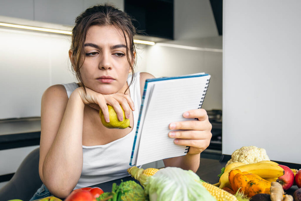 Молодая женщина на кухне с блокнотом среди овощей, концепция приготовления пищи, диеты и здорового питания. - Фото, изображение