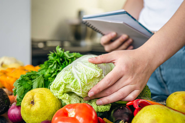 Mujer con bloc de notas y verduras en la mesa de la cocina, cocinar una receta, el concepto de dieta, contar calorías y perder peso. - Foto, imagen
