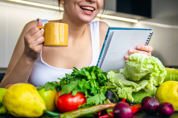 Una giovane donna in cucina con un blocco note tra le verdure, il concetto di cucina, dieta e alimentazione sana. - Foto, immagini
