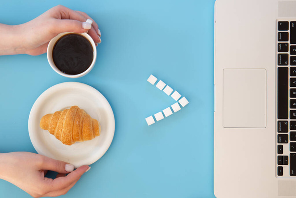 Croissant, koffiebeker en laptop op blauwe achtergrond, platte lay, conceptueel minimalisme. - Foto, afbeelding