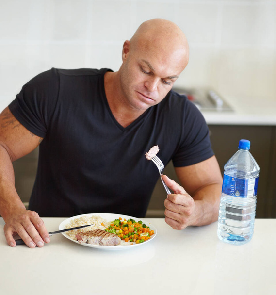 Πιέζοντας κάτω τελευταία μπουκιά του. Ένας μυώδης άντρας τρώει ένα ισορροπημένο γεύμα στον πάγκο της κουζίνας του και δείχνει γεμάτος. - Φωτογραφία, εικόνα
