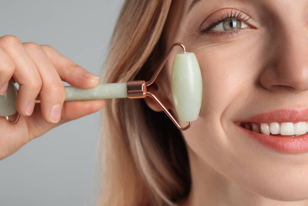Young woman using natural jade face roller on light grey background, closeup - Foto, afbeelding