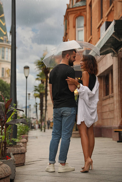 Νεαρό ζευγάρι με ομπρέλα απολαμβάνει το χρόνο μαζί κάτω από τη βροχή στο δρόμο της πόλης - Φωτογραφία, εικόνα