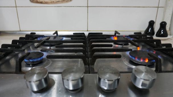 A blue fire is burning on the gas stove. Gas at home. Metal gas stove. Black bars. Light switches of burners. The gas is working. Beige tiles on the walls. Stop the economic crisis. Warmth in house - Photo, Image