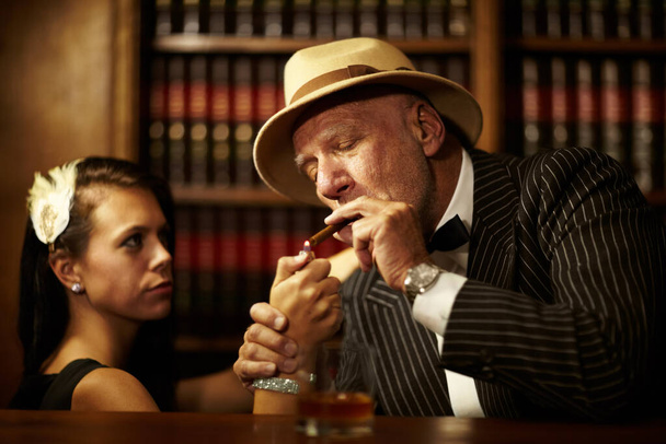 Έχει τον έλεγχο όλων γύρω του. Ένας ηλικιωμένος μαφιόζος που φοράει καπέλο και δείχνει σοβαρός ενώ μια γυναίκα ανάβει ένα τσιγάρο γι 'αυτόν. - Φωτογραφία, εικόνα