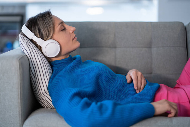 Νεαρή γυναίκα που φοράει ασύρματα ακουστικά και μπλε πουλόβερ βρίσκεται σε άνετο καναπέ και απολαμβάνει να ακούει ήρεμη μουσική. Γυναίκα ξεκουράζεται από κουραστική μέρα εργασίας ως ελεύθερος επαγγελματίας στο κοντινό γραφείο στο σπίτι - Φωτογραφία, εικόνα