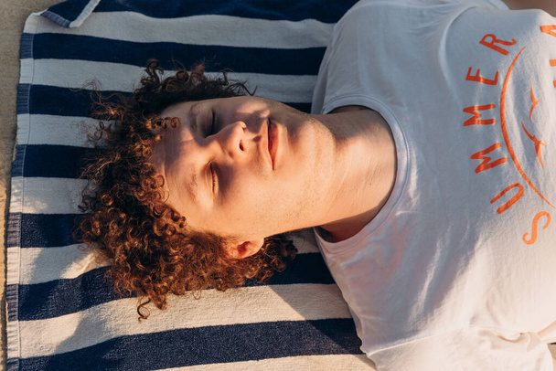 Jonge blanke man met krullend haar, gesloten ogen, rusten, ontspannen of slapen in wit T-shirt met woord Zomer ligt op gestreepte handdoek op het strand bij zonsondergang. Portret, vooraanzicht. - Foto, afbeelding
