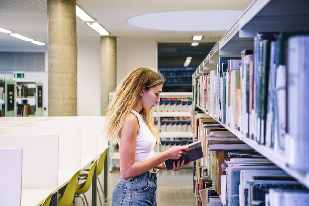 Πλευρική άποψη της νεαρής γυναίκας σε casual ρούχα με μακρύ βιβλίο ανάγνωσης μαλλιών, ενώ στέκεται κοντά σε βιβλιοθήκες στη βιβλιοθήκη του πανεπιστημίου - Φωτογραφία, εικόνα