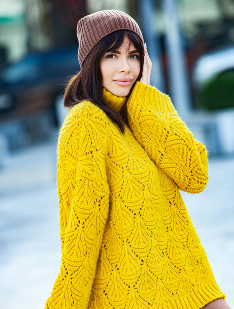 Beautiful European brunette dressed in a yellow woolen sweater and hat outside. The beautiful girl who wears thick stylish autumn clothes in cool weather. - Fotoğraf, Görsel