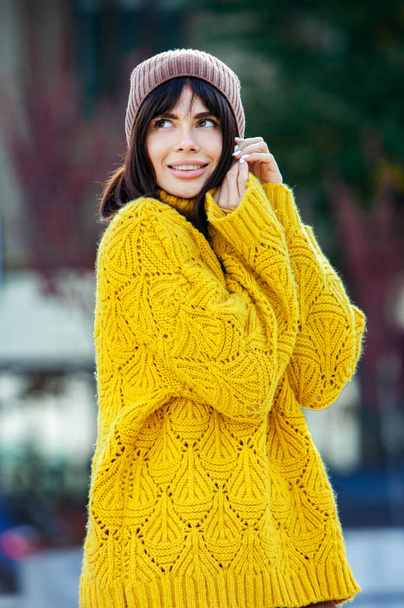 Schöne europäische Brünette in einem gelben Wollpullover und Hut draußen gekleidet. Das schöne Mädchen, das bei kühlem Wetter dicke, stylische Herbstkleidung trägt. - Foto, Bild