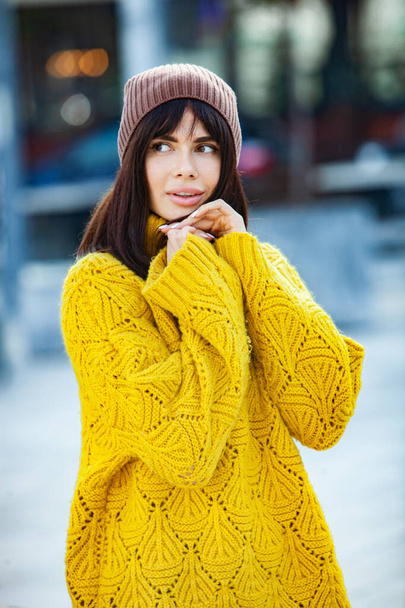 Beautiful European brunette dressed in a yellow woolen sweater and hat outside. The beautiful girl who wears thick stylish autumn clothes in cool weather. - Fotó, kép
