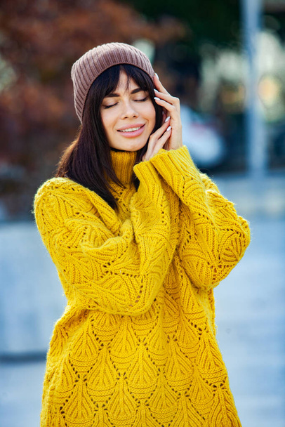 Beautiful European brunette dressed in a yellow woolen sweater and hat outside. The beautiful girl who wears thick stylish autumn clothes in cool weather. - Photo, Image