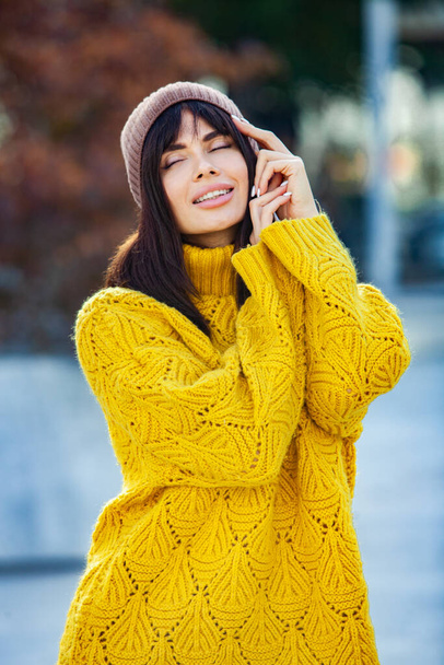 Piękna europejska brunetka ubrana w żółty wełniany sweter i kapelusz na zewnątrz. Piękna dziewczyna, która nosi grube stylowe jesienne ubrania w chłodną pogodę. - Zdjęcie, obraz