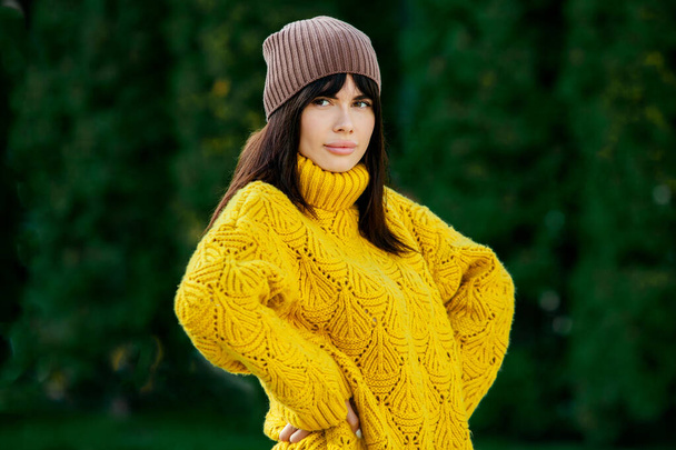 美しいヨーロッパのブルネットは黄色のウールのセーターと帽子を外で着ていました。涼しい天気の中で太いスタイリッシュな秋の服を身に着けている美しい女の子. - 写真・画像