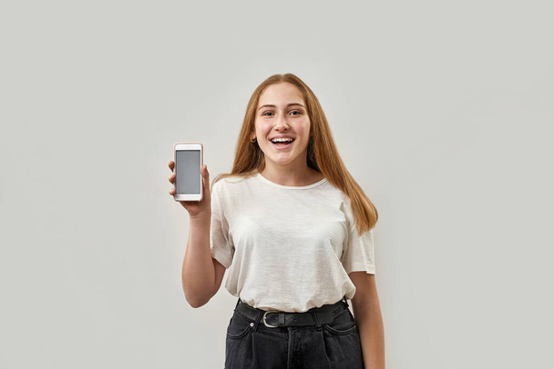 Smiling caucasian female teenage showing mobile phone and looking at camera. Girl with freckles of zoomer generation. Modern youngster lifestyle. Isolated on white background. Studio shoot. Copy space - Photo, Image