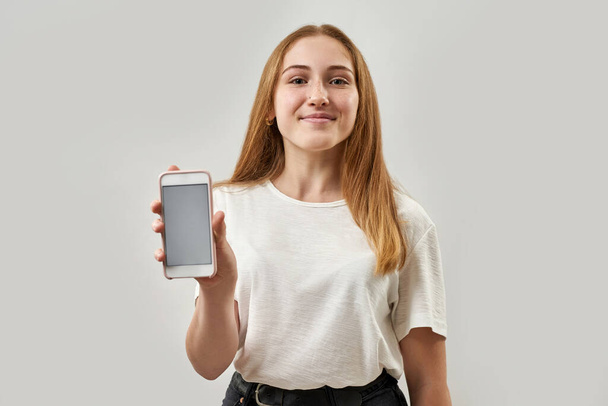 Front view of smiling caucasian female teenage holding and showing smartphone. Girl with freckles of zoomer generation. Modern youngster lifestyle. Isolated on white background in studio. Copy space - Photo, Image