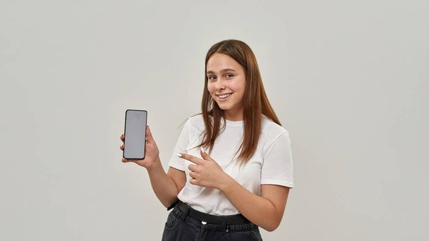 Smiling caucasian female teenage showing smartphone and looking at camera. Girl of zoomer generation. Concept of modern youngster lifestyle. Isolated on white background. Studio shoot. Copy space - Φωτογραφία, εικόνα