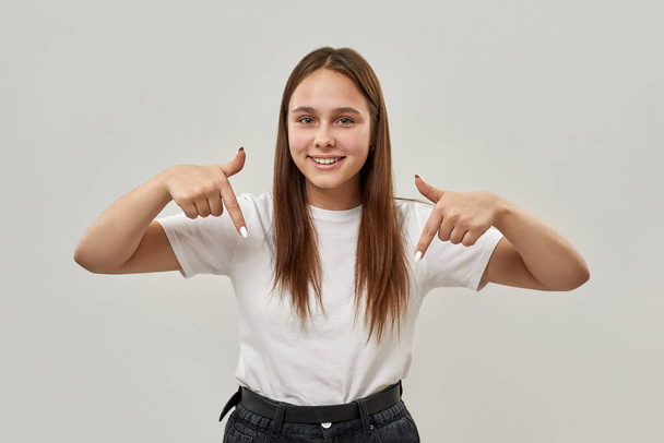 Smiling caucasian female teenage pointing with fingers down and looking at camera. Girl of zoomer generation. Concept of modern youngster lifestyle. Isolated on white background in studio. Copy space - Photo, Image