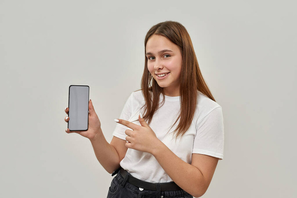 Smiling caucasian female teenage pointing with finger at smartphone and looking at camera. Girl of zoomer generation. Modern youngster lifestyle. Isolated on white background. Studio shoot. Copy space - Photo, image