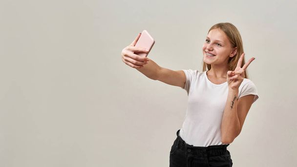 Sorridente ragazza caucasica adolescente che mostra segno di vittoria durante la presa selfie su smartphone. Bionda femmina bambina di generazione zoomer. Stile di vita giovanile moderno. Sfondo bianco. Scatto in studio. Copia spazio - Foto, immagini