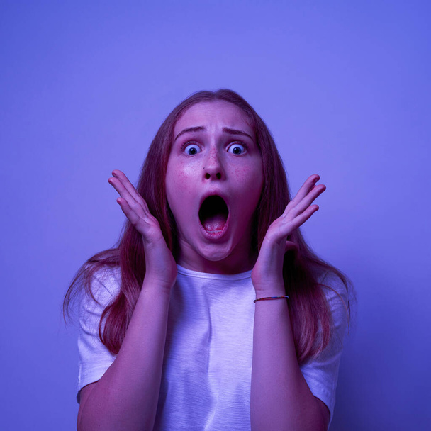 Scared caucasian female teenage with open mouth looking at camera. Excited girl of zoomer generation. Concept of modern youngster lifestyle. Isolated on blue with white light background. Studio shoot - Photo, Image