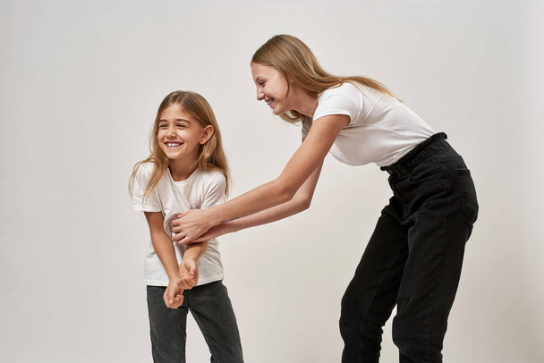 Teenage girl tickling her little sister. Joking blonde caucasian sisters of zoomer generation enjoying time together. Modern youngster lifestyle. Isolated on white background. Studio shoot. Copy space - Фото, изображение