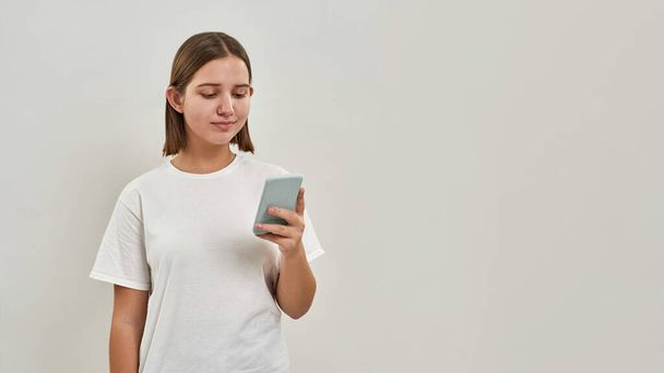 Concentrated caucasian teenage girl browsing smartphone. Female of zoomer generation wearing t-shirt. Concept of modern youngster lifestyle. Isolated on white background. Studio shoot. Copy space - Photo, Image