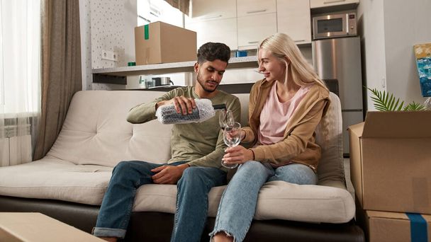 Middle eastern man pouring wine from bottle in glasses in hands of smiling blonde caucasian girl on sofa at modern spacious flat. Young multiracial couple celebrating moved to their new apartment - Fotó, kép