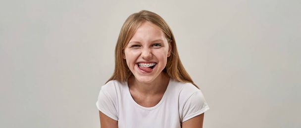 Portrait of laughing caucasian teenage girl sticking out tongue and looking at camera. Blonde female child of zoomer generation. Modern youngster lifestyle. White background. Studio shoot. Copy space - 写真・画像