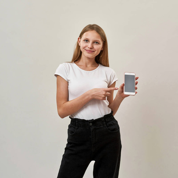 Front view of smiling caucasian teenage girl showing smartphone and looking at camera. Female child of zoomer generation. Modern youngster lifestyle. Isolated on white background in studio. Copy space - Foto, imagen