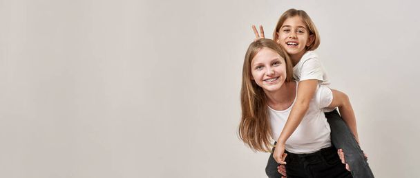 Teenage girl holding little sister piggyback while she making horns sign over her head. Joking caucasian sisters of zoomer generation looking at camera. White background. Studio shoot. Copy space - 写真・画像