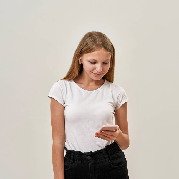 Focused caucasian teenage girl wearing earphones using smartphone. Beautiful blonde female child of zoomer generation. Modern youngster lifestyle. Isolated on white background in studio. Copy space - Φωτογραφία, εικόνα