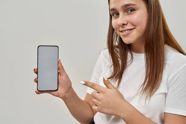 Cropped image of smiling european female teenage showing smartphone and looking at camera. Girl of zoomer generation. Modern youngster lifestyle. Isolated on white background. Studio shoot. Copy space - Photo, Image