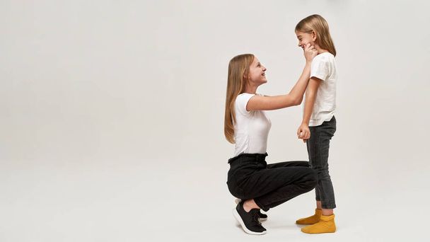 Side view of teenage girl tugging cheeks of little sister. joking caucasian sisters of zoomer generation looking at each other. Modern youngster lifestyle. White background. Studio shoot. Copy space - Photo, Image