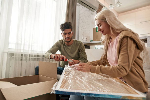 Young multiracial couple unpacking picture from stretch film with knife on sofa in modern spacious flat. Blonde caucasian girl and middle eastern man moved to their new apartment. Home relocation - Foto, Imagen