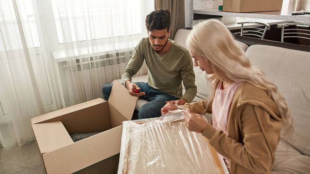 Young focused multiethnic couple unpacking picture from stretch film with knife on sofa in modern flat. Caucasian girl and middle eastern man moved to their new apartment. Home relocation - Zdjęcie, obraz