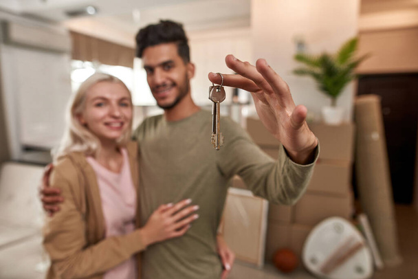 Selective focus of keys in hand of man hugging girl with background of cardboard boxes and things in new flat. Blurred young couple looking at camera. People moving to new apartment. Home relocation - Fotografie, Obrázek