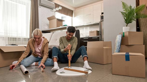 Concentrated caucasian girl and middle eastern man packing table on floor. Young multiethnic couple moving to their new apartment. Home relocation. Interior of modern spacious flat - Foto, Imagem