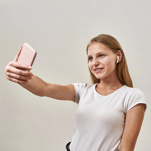 Partial of smiling caucasian teenage girl taking selfie on smartphone. Female child of zoomer generation wear earphones. Modern youngster lifestyle. Isolated on white background in studio. Copy space - Φωτογραφία, εικόνα