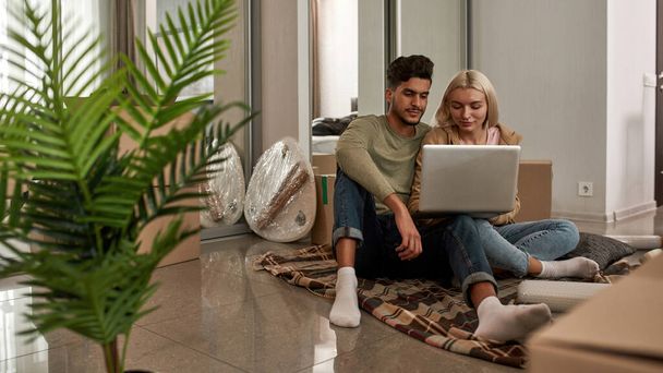 Young focused multiracial couple using and watching laptop on blanket on floor with cardboard boxes and things. Caucasian girl and middle eastern man move to new apartment. Home relocation - Fotó, kép
