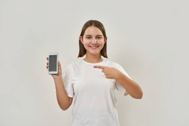 Smiling caucasian teenage girl pointing with finger at smartphone and looking at camera. Female of zoomer generation. Modern youngster lifestyle. Isolated on white background. Studio shoot. Copy space - Photo, image