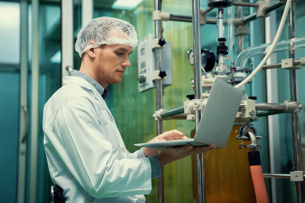 Apotekární vědec pracující s notebookem poblíž extraktoru oleje CBD a vědeckým strojem používaným k výrobě léčivých konopných výrobků. - Fotografie, Obrázek