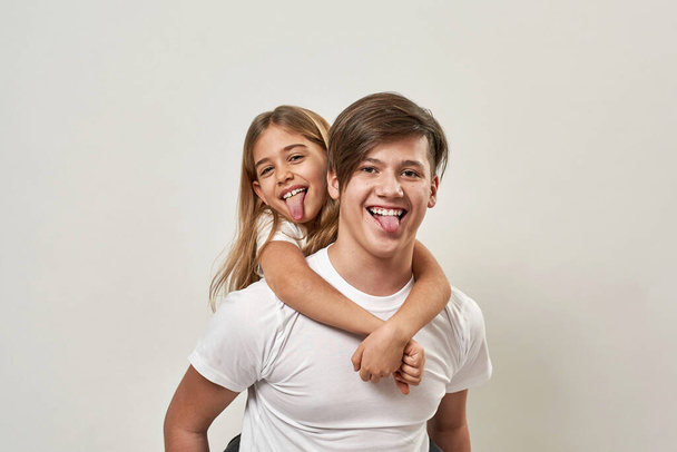 Teenage boy hold little sister piggyback and they stick out tongues. Joking caucasian guy and girl of zoomer generation look at camera. Youngster lifestyle. White background. Studio shoot. Copy space - Фото, зображення