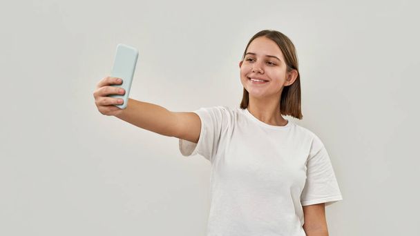 Smiling caucasian teenage girl taking selfie on mobile phone. Female of zoomer generation wearing t-shirt. Concept of modern youngster lifestyle. Isolated on white background. Studio shoot. Copy space - Φωτογραφία, εικόνα