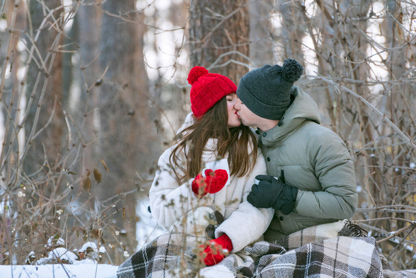 Young couple are sitting hugging and kissing in a snow-covered park, wrapping themselves in a plaid. Man and woman on a picnic in the winter forest - Photo, Image
