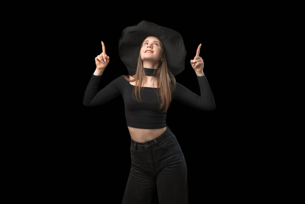 Meisje met puntige hoed en zwarte kleren wijst met de vingers omhoog. Portret van jonge heks is geïsoleerd op zwarte achtergrond. Kopieerruimte - Foto, afbeelding